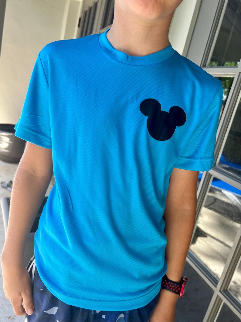 May include: A bright blue t-shirt featuring a black Mickey Mouse silhouette on the chest. The short-sleeved shirt is made of a lightweight, possibly athletic material. The shirt is a solid color with a crew neck.