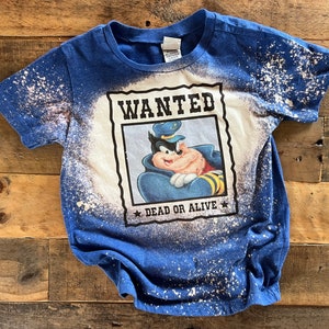 May include: A blue t-shirt with a white bleach splattered design. The shirt features a cartoon character in a police uniform with the text "WANTED DEAD OR ALIVE" on a poster.