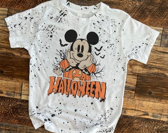Puede incluir: Camiseta blanca con un diseño de pintura salpicada negra. La camiseta presenta una imagen de dibujos animados de Mickey Mouse vestido de calabaza con la palabra "HALLOWEEN" impresa debajo.