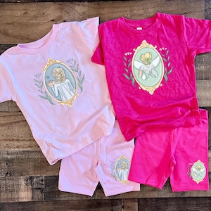 Peut inclure: Deux ensembles de t-shirt et short pour enfants. Un ensemble est rose avec une fille tenant une fleur dans un cadre dor&eacute;. L'autre ensemble est rose avec une f&eacute;e dans un cadre dor&eacute;.