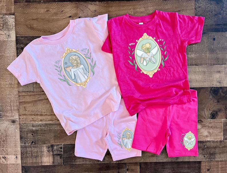 Peut inclure: Deux ensembles de t-shirt et short pour enfants. Un ensemble est rose avec une fille tenant une fleur dans un cadre dor&eacute;. L'autre ensemble est rose avec une f&eacute;e dans un cadre dor&eacute;.
