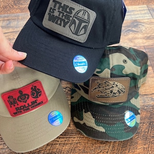 May include: Several baseball caps with Star Wars designs. A black cap displays "THIS IS THE WAY" on a patch. A tan cap has a red patch with "ROLLIN' WITH THE HOMIES". A camouflage cap features a Millennium Falcon patch.