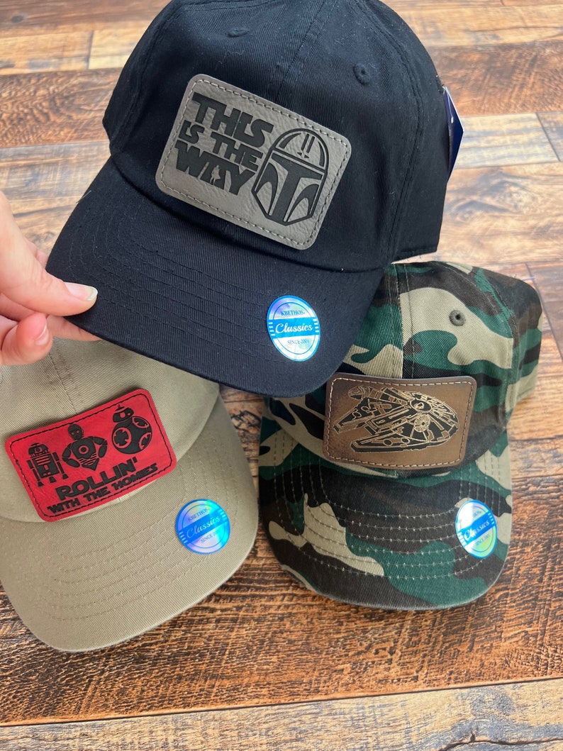 May include: Several baseball caps with Star Wars designs. A black cap displays "THIS IS THE WAY" on a patch. A tan cap has a red patch with "ROLLIN' WITH THE HOMIES". A camouflage cap features a Millennium Falcon patch.