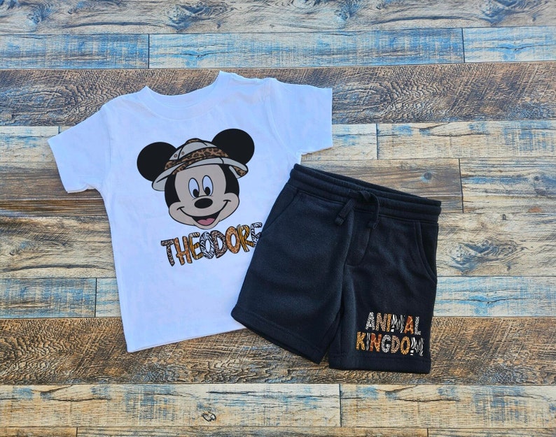 May include: White t-shirt with a Mickey Mouse character wearing a safari hat and the text "Theodore". Black sweatpants with the text "Animal Kingdom".