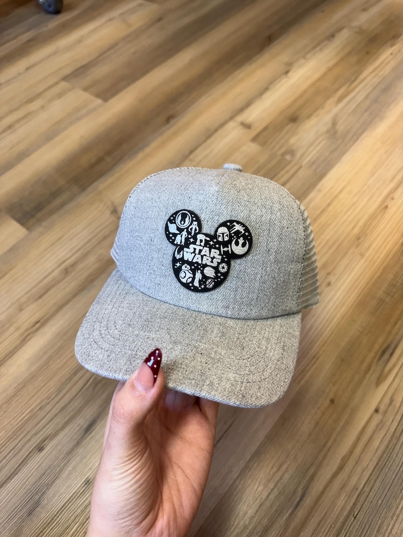 May include: A grey Star Wars baseball cap with a mesh back and a curved peak. The front has a Mickey Mouse head design with Star Wars characters and logos. The cap is made of a textured fabric.
