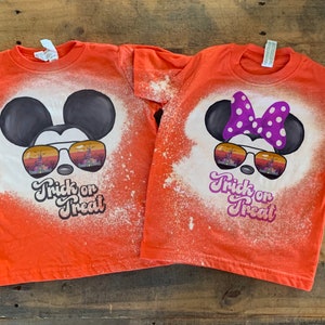 Puede incluir: Dos camisetas naranjas con un dise&ntilde;o salpicado de lej&iacute;a que presenta a Mickey Mouse y Minnie Mouse con gafas de sol. El texto "Trick or Treat" est&aacute; impreso en ambas camisetas.