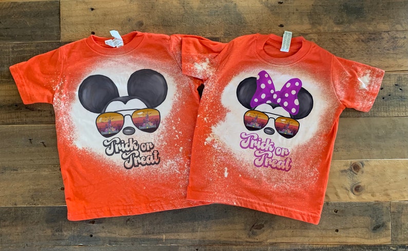Puede incluir: Dos camisetas naranjas con un dise&ntilde;o salpicado de lej&iacute;a que presenta a Mickey Mouse y Minnie Mouse con gafas de sol. El texto "Trick or Treat" est&aacute; impreso en ambas camisetas.
