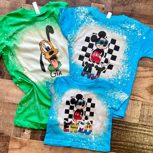 May include: Three bleached t-shirts with cartoon character designs. The green shirt has a Goofy design with the name "GIA". The blue shirt has a Mickey Mouse design with the name "AJ". The smaller blue shirt has a Mickey Mouse design with the name "ENZO".