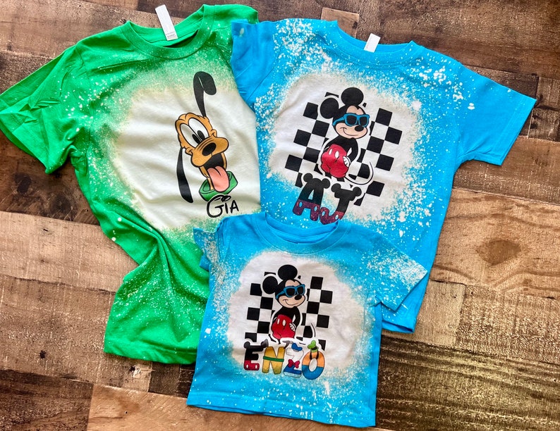 May include: Three bleached t-shirts with cartoon character designs. The green shirt has a Goofy design with the name "GIA". The blue shirt has a Mickey Mouse design with the name "AJ". The smaller blue shirt has a Mickey Mouse design with the name "ENZO".