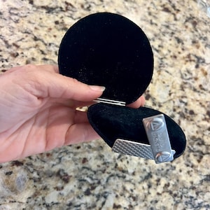 K&ouml;nnte beinhalten: Eine schwarze Samt-Schmuckschatulle mit silbernem Verschluss, ge&ouml;ffnet, um eine silberne Krawattenklammer freizugeben. Die Krawattenklammer hat ein strukturiertes Design und den Text "DAVID MAGNETS".