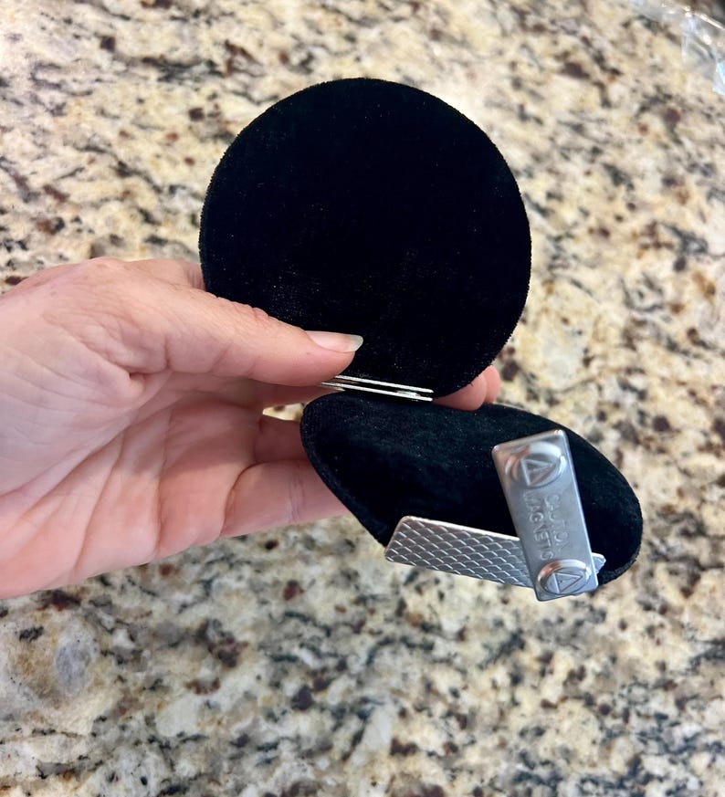 K&ouml;nnte beinhalten: Eine schwarze Samt-Schmuckschatulle mit silbernem Verschluss, ge&ouml;ffnet, um eine silberne Krawattenklammer freizugeben. Die Krawattenklammer hat ein strukturiertes Design und den Text "DAVID MAGNETS".