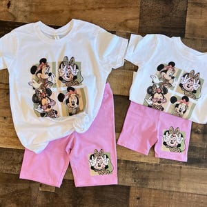 May include: A white t-shirt with a pink pair of shorts. The t-shirt has a graphic print of four different images of Mickey Mouse with a leopard print design. The shorts have a single image of Mickey Mouse with a leopard print design on the left leg.