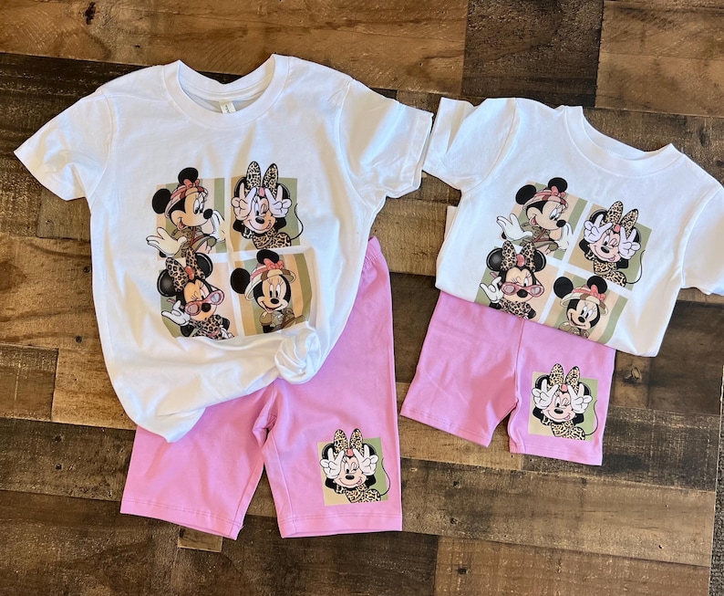 May include: A white t-shirt with a pink pair of shorts. The t-shirt has a graphic print of four different images of Mickey Mouse with a leopard print design. The shorts have a single image of Mickey Mouse with a leopard print design on the left leg.