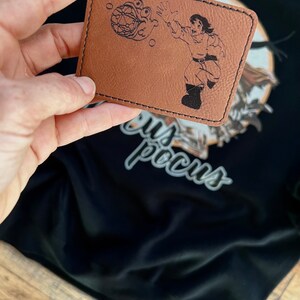 May include: A brown leather patch with a black line drawing of a person holding a sphere with a face on it. The text "Hocus Pocus" is visible in the background.