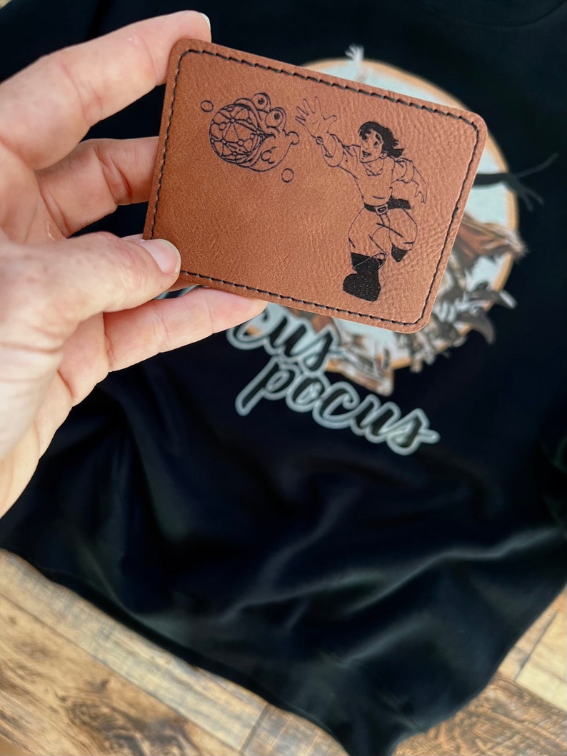 May include: A brown leather patch with a black line drawing of a person holding a sphere with a face on it. The text "Hocus Pocus" is visible in the background.