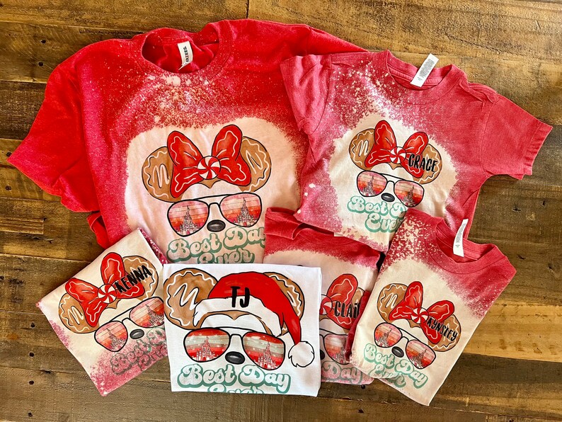 May include: Set of five red, white, and green Christmas themed t-shirts with a gingerbread Mickey Mouse design. The shirts have the text "Best Day Ever" and personalized names on each shirt.