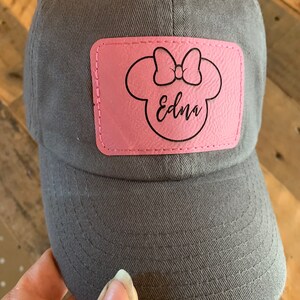 Pu&ograve; includere: Un cappello da baseball grigio con una toppa in pelle rosa che presenta una silhouette nera di una testa di topo con un fiocco e il nome "Edna" scritto in corsivo.