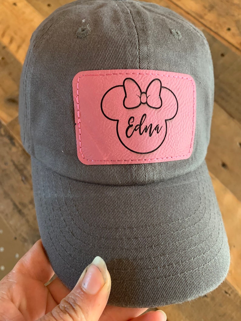 Pu&ograve; includere: Un cappello da baseball grigio con una toppa in pelle rosa che presenta una silhouette nera di una testa di topo con un fiocco e il nome "Edna" scritto in corsivo.