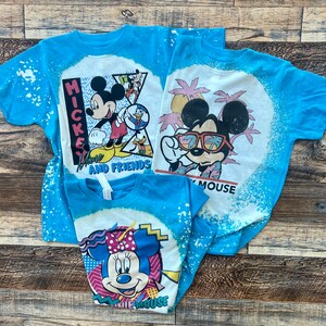 Puede incluir: Tres camisetas azules con dise&ntilde;os de decoloraci&oacute;n con lej&iacute;a que representan a Mickey Mouse y Minnie Mouse. Las camisetas tienen dise&ntilde;os diferentes con el texto "Have a Mouse and Friends" y "Have a Mouse".