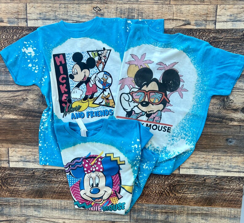 Puede incluir: Tres camisetas azules con dise&ntilde;os de decoloraci&oacute;n con lej&iacute;a que representan a Mickey Mouse y Minnie Mouse. Las camisetas tienen dise&ntilde;os diferentes con el texto "Have a Mouse and Friends" y "Have a Mouse".