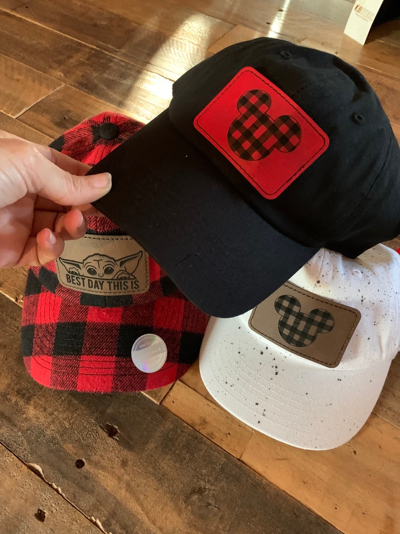 May include: Three baseball caps with different designs. One black cap with a red and black plaid Mickey Mouse patch, one red and black plaid cap with a "Best Day This Is" patch, and one white cap with a brown and black plaid Mickey Mouse patch.