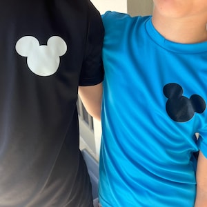 May include: Two short-sleeved shirts, one black and one blue, each featuring a white or black Mickey Mouse silhouette on the upper left chest. The shirts appear to be made of a lightweight, athletic material.