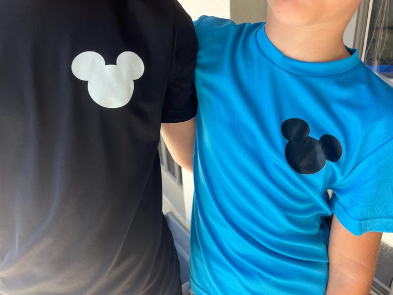 May include: Two short-sleeved shirts, one black and one blue, each featuring a white or black Mickey Mouse silhouette on the upper left chest. The shirts appear to be made of a lightweight, athletic material.
