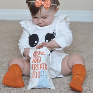May include: A baby wearing a white ghost costume with a black face and eyelashes, orange socks, and an orange glitter headband. The baby is holding a white treat bag with the text "Babies Need Treats Too" and a blue ghost illustration.