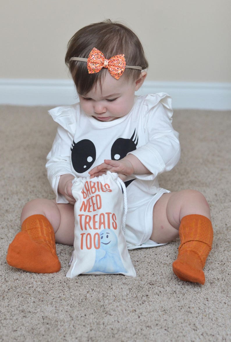 May include: A baby wearing a white ghost costume with a black face and eyelashes, orange socks, and an orange glitter headband. The baby is holding a white treat bag with the text "Babies Need Treats Too" and a blue ghost illustration.