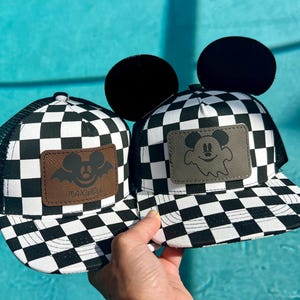 May include: Two black and white checkered trucker hats with mesh backs. One hat features a brown leather patch with a bat-eared mouse and the name Maxwell. The other hat has a grey leather patch with a ghost-shaped mouse and black mouse ears.
