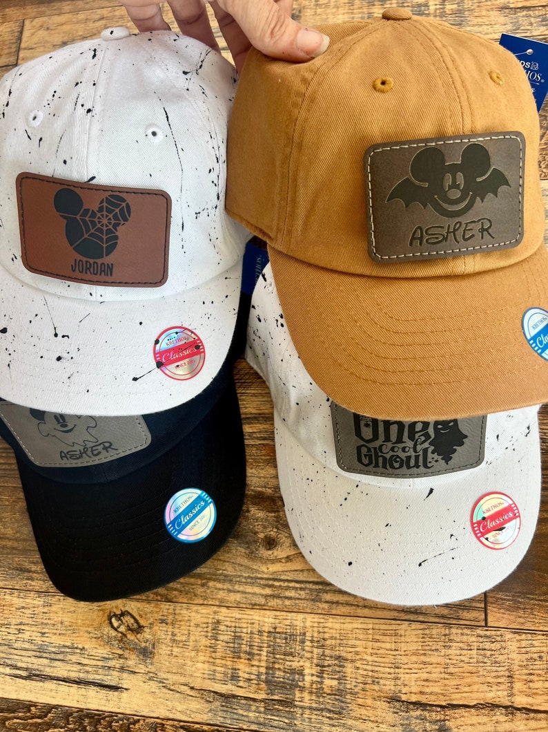 May include: Four baseball caps with black and white splatter paint designs. Each cap has a personalized leather patch with a Mickey Mouse design and a name. The names on the patches are Jordan, Asher, One Cool Ghoul, and Asher.
