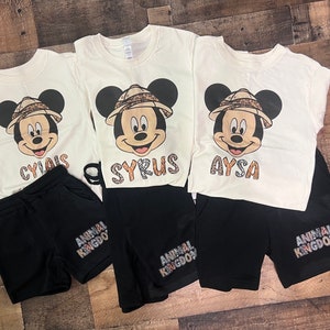 May include: Three white t-shirts with a cartoon image of Mickey Mouse wearing a safari hat and a name printed below. Each shirt is paired with black shorts that have "Animal Kingdom" printed on them.