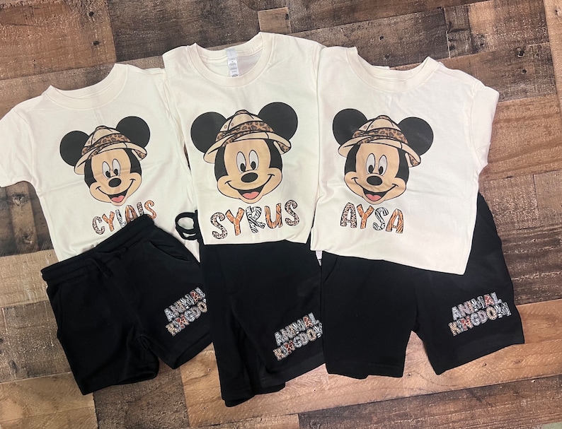 May include: Three white t-shirts with a cartoon image of Mickey Mouse wearing a safari hat and a name printed below. Each shirt is paired with black shorts that have "Animal Kingdom" printed on them.