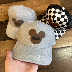 May include: Three grey trucker caps with personalised leather patches. One cap has a black and white chequered pattern. The patches feature the names "MAX", "NOAH", and "RYDER" in a playful font, set against a brown Mickey Mouse silhouette.
