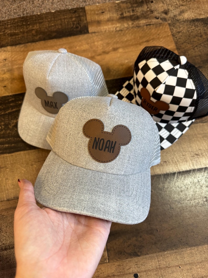 May include: Three grey trucker caps with personalised leather patches. One cap has a black and white chequered pattern. The patches feature the names "MAX", "NOAH", and "RYDER" in a playful font, set against a brown Mickey Mouse silhouette.