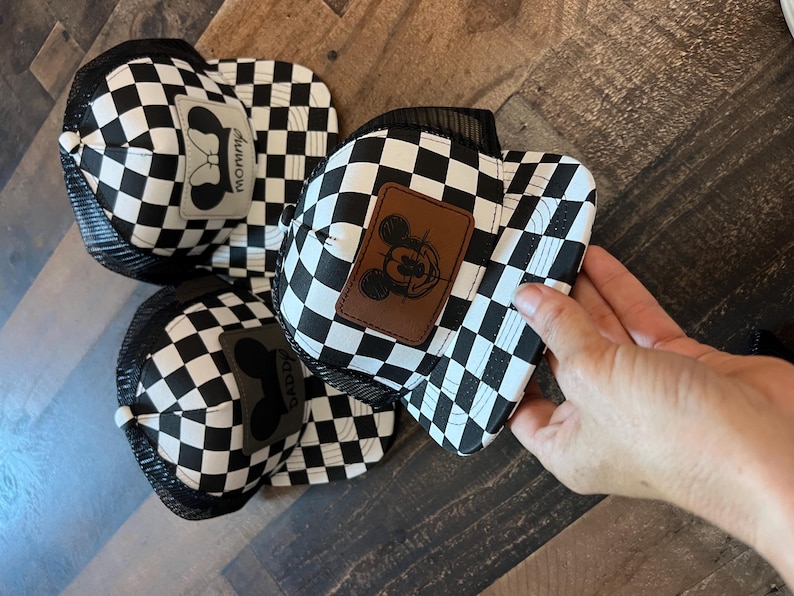 K&ouml;nnte beinhalten: Schwarz-wei&szlig; karierte Trucker-H&uuml;te mit Mesh-R&uuml;cken. Ein Hut hat einen braunen Aufn&auml;her mit einem Mickey-Mouse-Design, ein anderer hat einen grauen Aufn&auml;her mit dem Wort "Mommy". Der dritte Hut hat einen grauen Aufn&auml;her mit dem Wort "Daddy".