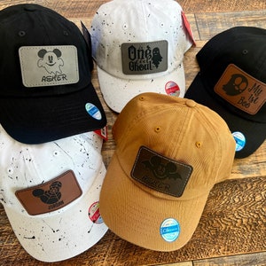 May include: Four baseball caps with different designs. The caps are black, white, brown, and white with black splatter paint. Each cap has a leather patch with a Mickey Mouse design and a name. The names on the patches are Asher, Jordan, One Cool Ghoul, and My 1st Boo.