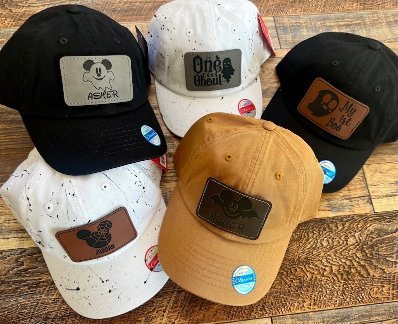 May include: Four baseball caps with different designs. The caps are black, white, brown, and white with black splatter paint. Each cap has a leather patch with a Mickey Mouse design and a name. The names on the patches are Asher, Jordan, One Cool Ghoul, and My 1st Boo.