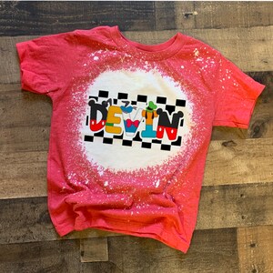 May include: Red t-shirt with a bleach-splattered design featuring a checkered background and the name "DEVIN" in white letters with Disney characters incorporated into the letters.