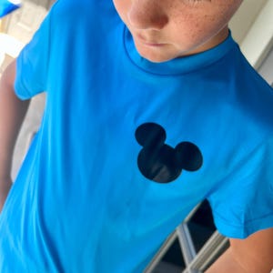 May include: Bright blue t-shirt featuring a black Mickey Mouse silhouette on the chest. The shirt has short sleeves and a crew neck. The fabric appears to be a lightweight, breathable material.