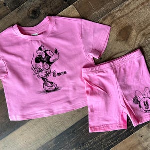 May include: Pink two-piece outfit for a child with a black and white graphic of Minnie Mouse on both the shirt and shorts. The shirt has the name "Emma" printed on it.