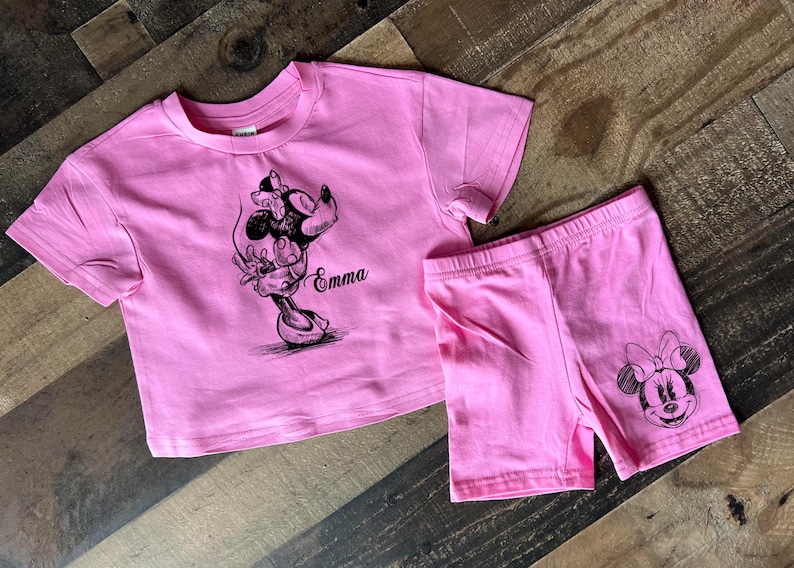 May include: Pink two-piece outfit for a child with a black and white graphic of Minnie Mouse on both the shirt and shorts. The shirt has the name "Emma" printed on it.
