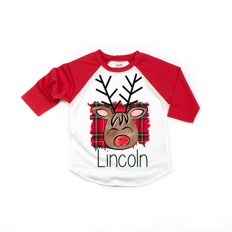 May include: White and red baseball tee shirt with a cartoon reindeer face on a plaid background. The text "Lincoln" is printed below the reindeer.