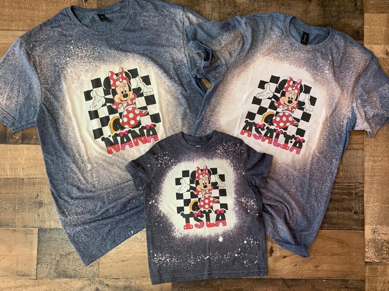 May include: Three blue, white, and red bleached t-shirts with a cartoon image of Minnie Mouse in a checkered pattern. The shirts are personalized with the names "Nana", "Asalta", and "Isla".