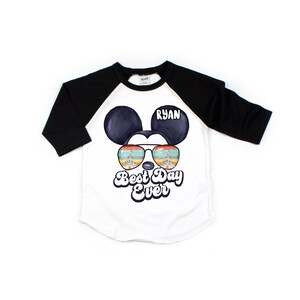 Peut inclure: T-shirt de baseball blanc et noir avec un dessin anim&eacute; de Mickey Mouse portant des lunettes de soleil avec un reflet de ch&acirc;teau. Le texte "Best Day Ever" est imprim&eacute; sous l'image. Le nom "Ryan" est imprim&eacute; sur le t-shirt.