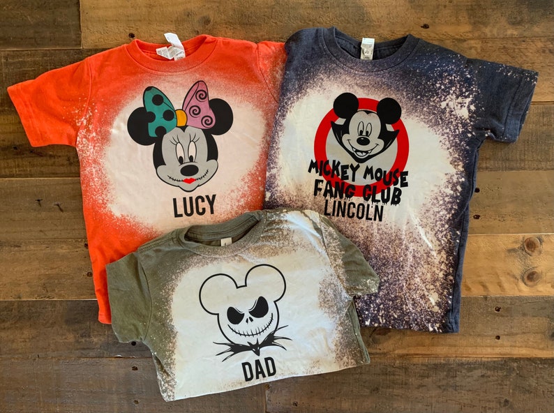 Puede incluir: Tres camisetas blanqueadas con diferentes dise&ntilde;os de tem&aacute;tica de Halloween. La primera camiseta es naranja con un dise&ntilde;o de Mickey Mouse en blanco y negro y el texto "LUCY". La segunda camiseta es azul marino con un c&iacute;rculo rojo alrededor de un dise&ntilde;o de Mickey Mouse en blanco y negro y el texto "MICKEY MOUSE FANG CLUB LINCOLN". La tercera camiseta es verde con un dise&ntilde;o de Jack Skellington en blanco y negro y el texto "DAD".