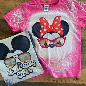 Puede incluir: Dos camisetas de algod&oacute;n blanqueadas con un dise&ntilde;o de Mickey Mouse y Minnie Mouse. La camiseta rosa presenta a Minnie Mouse con un lazo rojo y gafas de sol con una vista del Castillo de Cenicienta. La camiseta blanca presenta a Mickey Mouse con gafas de sol y el texto "Best Day Ever".