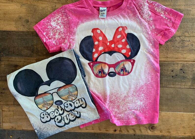 Puede incluir: Dos camisetas de algod&oacute;n blanqueadas con un dise&ntilde;o de Mickey Mouse y Minnie Mouse. La camiseta rosa presenta a Minnie Mouse con un lazo rojo y gafas de sol con una vista del Castillo de Cenicienta. La camiseta blanca presenta a Mickey Mouse con gafas de sol y el texto "Best Day Ever".