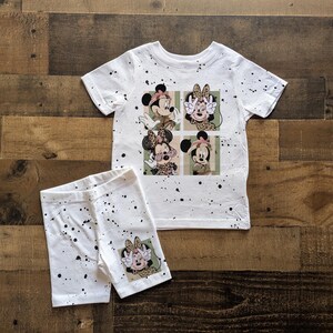 May include: White t-shirt with a splatter paint design featuring a cartoon image of four Mickey Mouse characters. The shirt is paired with white shorts with a similar splatter paint design.