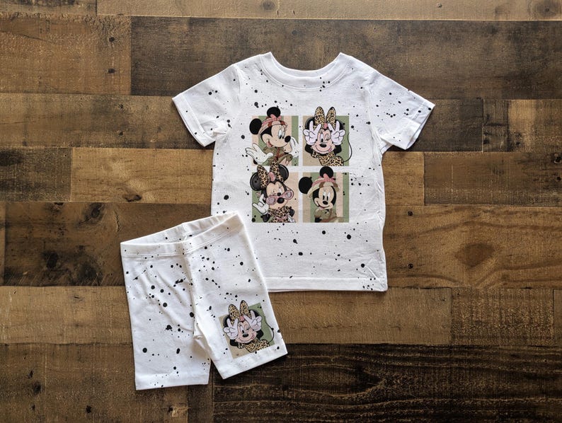 May include: White t-shirt with a splatter paint design featuring a cartoon image of four Mickey Mouse characters. The shirt is paired with white shorts with a similar splatter paint design.